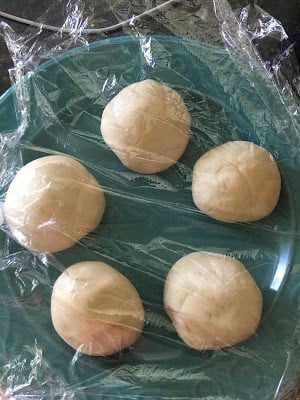 Five rounded smoothened portions of dough placed on a plate and covered with a plastic wrap.