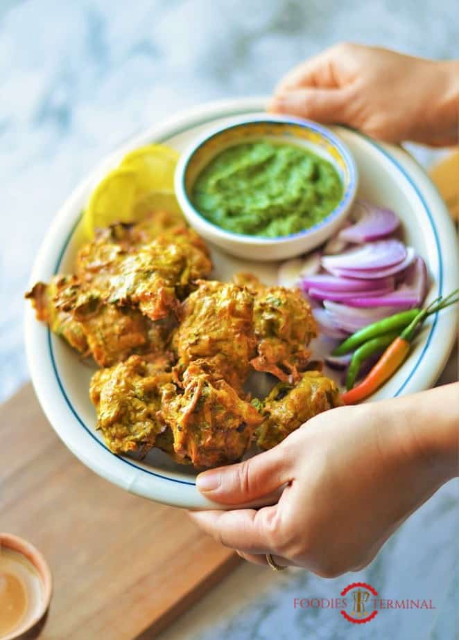 Chicken Pakora served in an oval white plate with chilies, onion slices, cilantro chuntey & lemon slices.