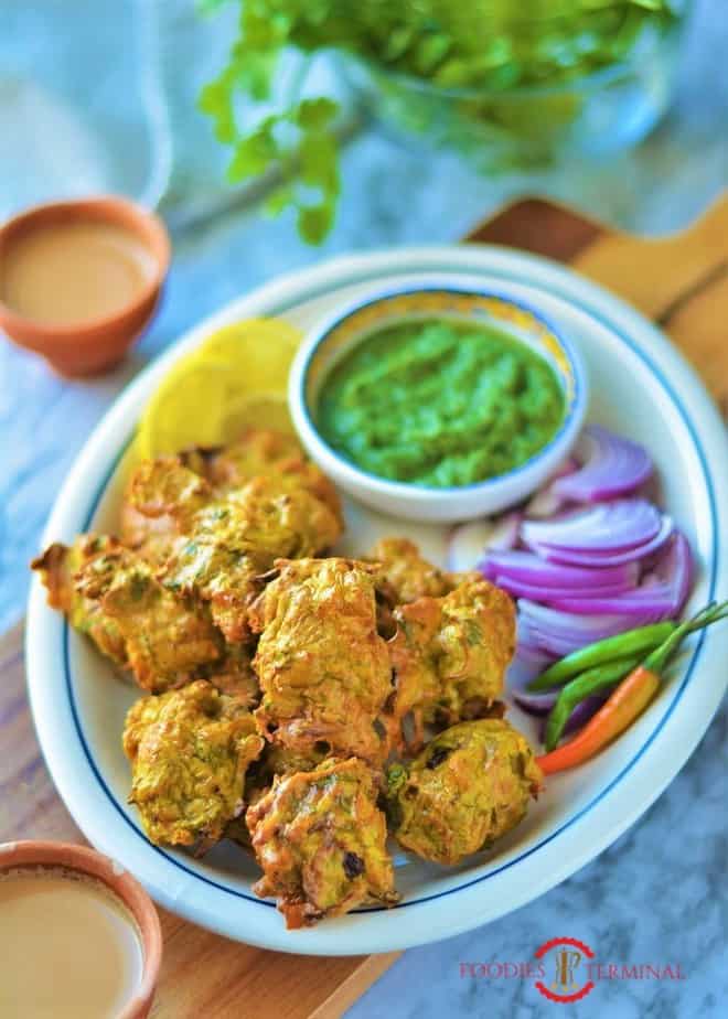 Chicken Pakora on a white oval plate with blue rim with onion slices, green chilies, green chutney