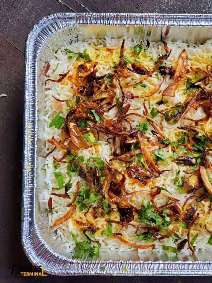 chicken Biryani topped with birista & cilantro mint leaves in an aluminum tray ready to go in oven