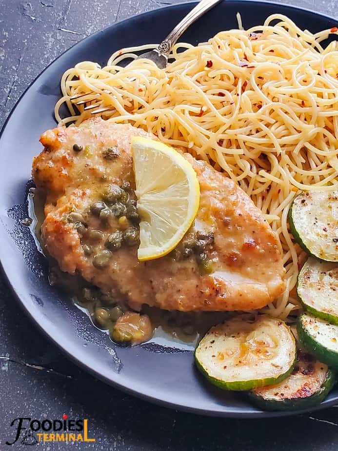 easy chicken piccata served with noodles & zucchini & a lemon wedge on a black plate