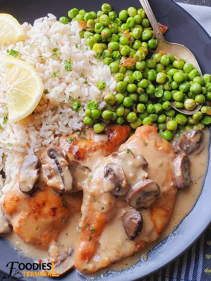 chicken marsala served on a black plate with brown rice, green peas & lemon wedges